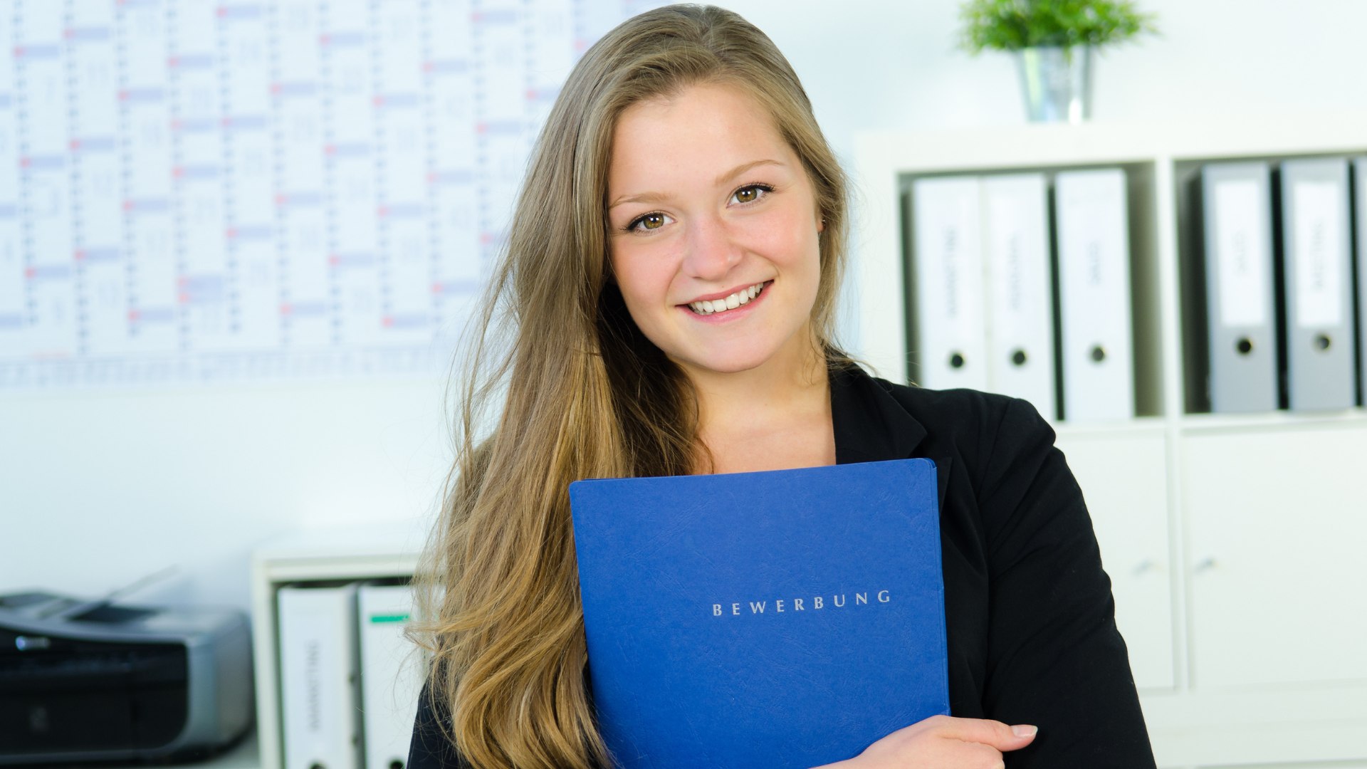Eine junge Dame steht vor einem Aktenschrank mit einer Bewerbungsmappe in der Hand und lächelt in die Kamera.