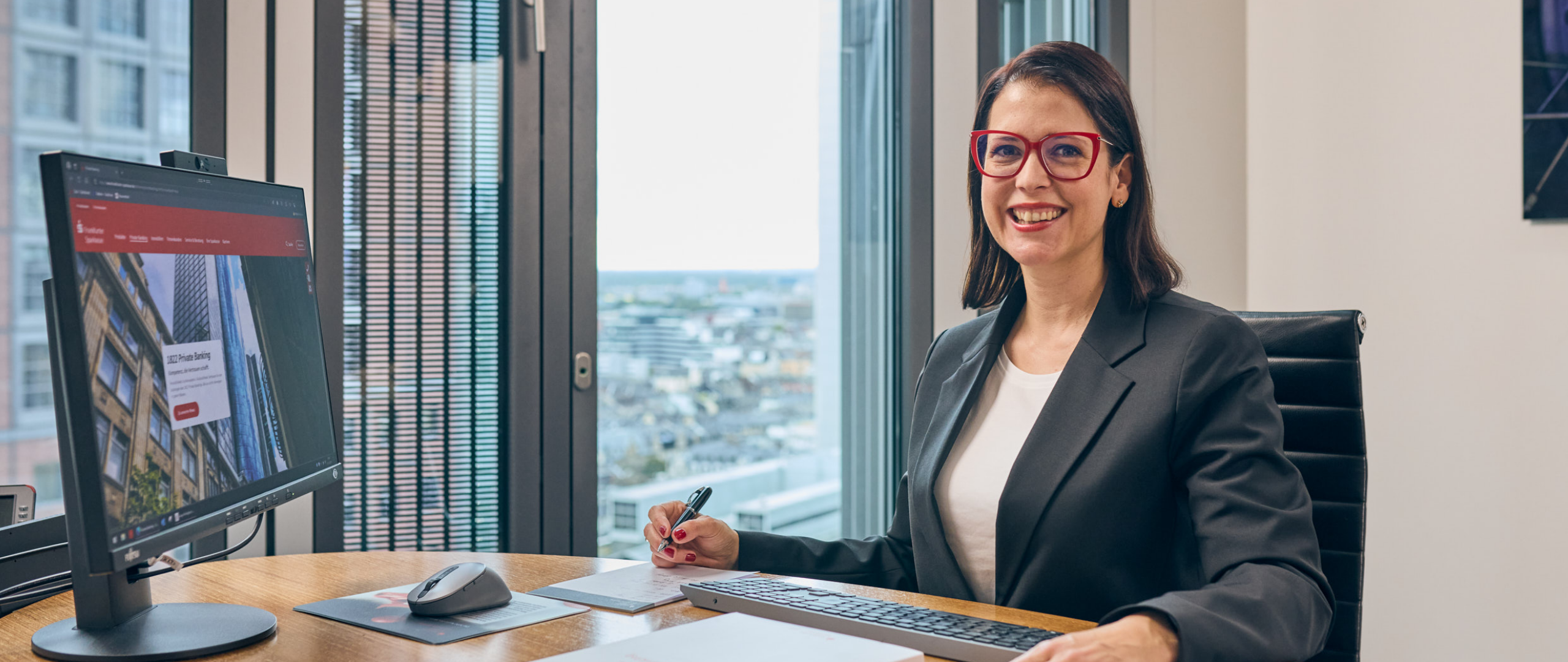 Private Banking Beraterin sitzt an ihrem Arbeitsplatz, Frankfurter Skyline im Hintergrund